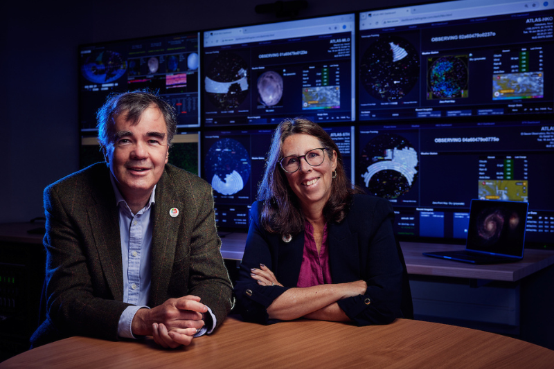 Richard and Julie Buchanan sitting side by side at a desk in the Astrophysics lab with screens displaying data in the background